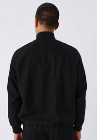 Giacca nera e strutturata con colletto alto, maniche raglan e orlo elasticizzato, vista da dietro. Nessun hardware o motivo visibile.