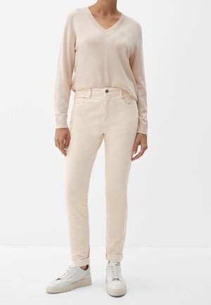 Beige v-aukkoinen neulepaita, pehmeä neulottu kangas; vaaleat, istuvat farkut, joissa viisi taskua ja kevyt jousto; yhdistetty valkoisiin lenkkareihin.