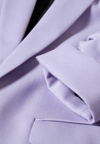 Gros plan d'une manche et d'une poche pliées d'un blazer violet clair montrant la texture du tissu et les détails de couture soignés.