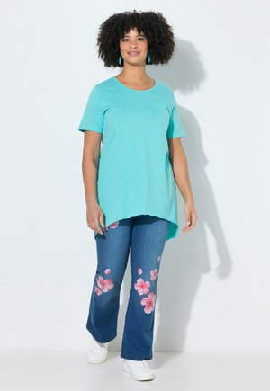 Woman with curly hair wearing turquoise short-sleeve shirt, blue jeans with pink flower prints, white sneakers, standing against white background.