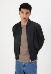 Giubbotto bomber nero con texture liscia, chiusura con zip e tasche laterali. Presenta una patch sulla manica e piccoli elementi decorativi.