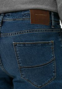 Jeans in denim blu scuro con una patta in pelle marrone riportante la scritta "MASTERPIECE" e "BPAX" in rilievo. Include tasche posteriori e cuciture a contrasto.