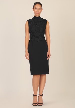 Abito nero con un corpino in pizzo floreale e una gonna aderente e slanciata. Presenta una scollatura drappeggiata e senza maniche, abbinato a tacchi neri.
