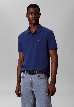 Mann trägt ein dunkelblaues Poloshirt mit kleinem Logo und hellblaue Jeans, steht vor grauem Hintergrund.