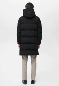 Abrigo acolchado negro con capucha, que presenta segmentos acolchados. Textura suave, diseño de longitud media, combinado con pantalones gris claros y zapatos negros.