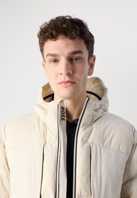 Jachetă puffer de culoare bej deschis, cu glugă, prevăzută cu închidere cu fermoar, accente negre și un logo pe guler. Textură netedă, căptușită.