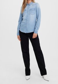 Camicia in denim azzurro chiaro a maniche lunghe, con chiusura a bottoni e due tasche sul petto, abbinata a pantaloni neri e scarpe con tacco bianche.