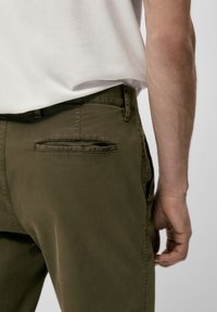 Olijfgroene broek van gladde stof, met een vlakke achterzak en een standaard riemlus, gecombineerd met een effen wit overhemd.