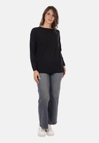 Donna in piedi che indossa una maglietta nera a maniche lunghe, jeans blu e sneakers bianche su uno sfondo bianco semplice.