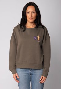 Brauner Baumwoll-Sweatshirt mit entspannter Passform, das ein grafisches Design und ein runder Aufnäher zeigt. Kombiniert mit blauen Jeans.