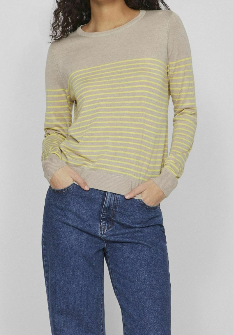 Pull beige à manches longues avec de fines rayures jaunes horizontales, porté avec un jean bleu, mains dans les poches, sur un fond uni.