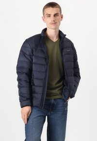 Tamsiai mėlynas pūkinis švarkas su užtrauktuku priekyje, turintis stovintį apykaklę, horizontalią siuvinėjimo struktūrą ir logotipo detalę kairėje krūtinėje.