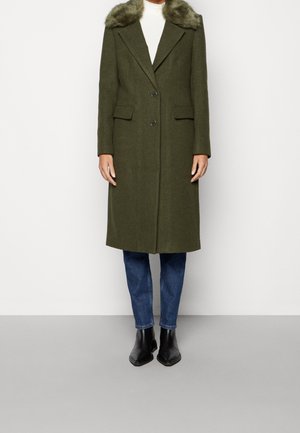 Manteau en laine vert olive avec col en fausse fourrure, fermeture à double bouton, deux poches avant et coupe droite, porté avec un jean bleu et des bottines noires.