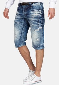 Shorts en denim dans un délavé bleu clair avec des détails déchirés, cinq poches et une coupe décontractée, associés à des baskets blanches.