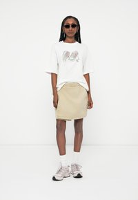 Biely oversized tričko s grafikou tenisky, béžová džínsová sukňa a sivé tenisky s čiernymi podrážkami. Modelka nosí tmavé slnečné okuliare a biele ponožky.