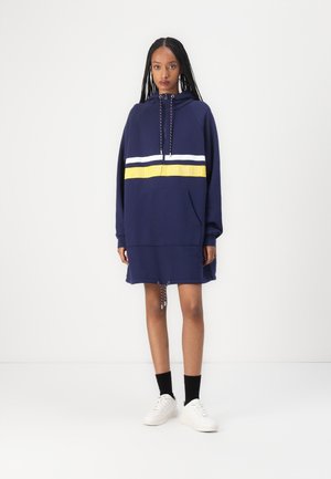 Navy blue hooded dress with white and yellow stripe, front pocket, adjustable drawstring, and textured fabric. Paired with white sneakers.
