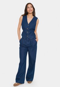 Džinsinė liemenė su V formos iškirpte, penkių rudos spalvos sagų užsegimu ir banguotu hemu. Suderinta su plačios kojos džinsais, abu tamsiai mėlynos spalvos.