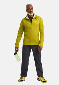 Lichtgroene zip-up jas met een witte kraag, grijze broek en zwarte wandelschoenen. Houdt een groene waterfles vast, staat voor een effen achtergrond.