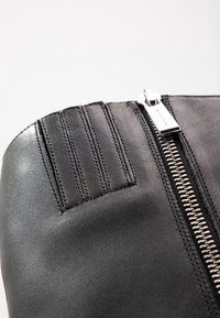 Detailní záběr na černou koženou botu, která ukazuje zip a čtyři vertikálně prošívané panely poblíž horního okraje.