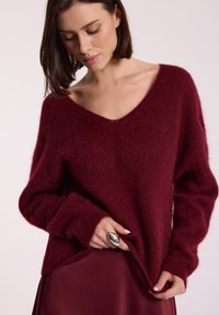 Burgundowy sweter z dzianiny z dekoltem w serek, w oversize'owym kroju, z ribbowanymi mankietami i miękką fakturą, w połączeniu z satynową burgundową spódnicą.