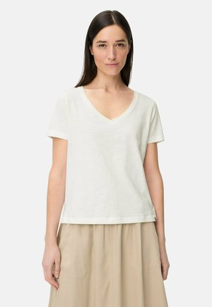 Kvinde med glat mørkt hår iført en hvid V-hals kortærmet skjorte og beige nederdel, stående foran en ensfarvet lys baggrund.