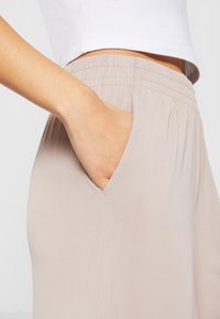 Main reposant dans la poche d'un pantalon beige à taille élastique porté avec un haut court blanc sur un fond clair.