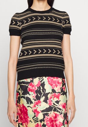 Kvinde iført en sort kortærmet strikbluse med beige geometriske striber og et sort skørt med et rødt og gult blomsterprint.
