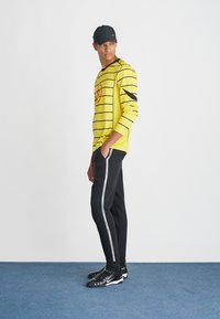 Kollane pikakäisteline särk mustade triipudega, kombineeritud mustade pükstega, millel on valge külge triip; must nokamüts ja mustad spordijalatsid täiendavad välimust.