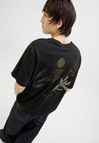 T-shirt en coton noir avec des illustrations de plantes et d'un soleil en art linéaire coloré au dos. Coupe décontractée avec des manches courtes.