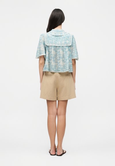 Γυναίκα με μακριά σκούρα μαλλιά, που φοράει γαλάζιο λουλουδάτο πουκάμισο με δαντελένιο τελείωμα και μπεζ σορτς, στέκεται μπροστά σε απλό λευκό φόντο.