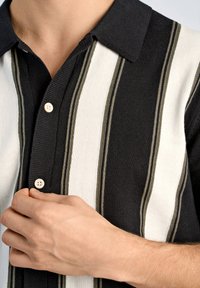 Svartvit randig kortärmad pikétröja med en strukturerad stickning. Har en spetsig krage och ljusa knappar.