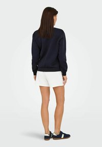 Marineblå sweatshirt med ribstrikkede manchetter og kant, parret med hvide shorts og marineblå sko, set fra bagen. Glat stoftekstur.