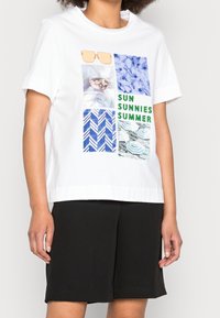 Camiseta blanca de manga corta con un diseño gráfico que presenta patrones en azul y verde, una imagen de gafas de sol y el texto "SUN SUNNIES SUMMER."