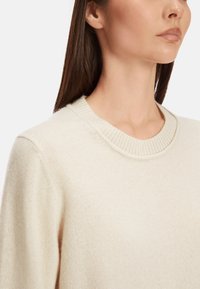 Hellcremefarbener Pullover mit geripptem Rundhalsausschnitt, weichem Material und lässiger Passform, der subtile Stickdetails präsentiert.
