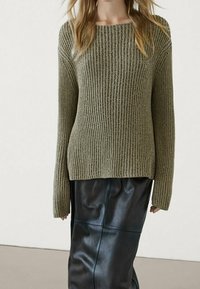 Pull en tricot vert olive avec une texture côtelée et un large décolleté, associé à une jupe mi-longue en cuir noir avec des détails en panneaux.