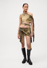 Dvojdielny outfit pozostávajúci z topu s jedným ramenom a zavinovacích sukne z patterned, ľahkej tkaniny. Farby zahŕňajú žltú, šedú a čiernu. Čierne vysoké čižmy po kolená.
