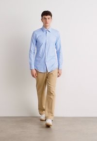 Camisa de manga longa azul clara com colarinho abotoado, combinada com calças bege e ténis brancos, em pé contra um fundo neutro.
