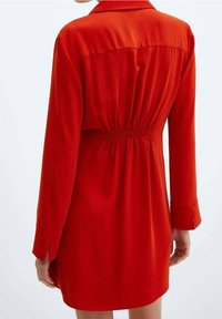 Vestido rojo con mangas largas, cuello y cintura elástica. Tejido suave; los fruncidos en la parte posterior crean textura. Aberturas laterales en los puños de las mangas.