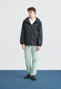 Νεαρός άνδρας που φοράει μαύρο μπουφάν με κουκούλα, λευκό μπλουζάκι, ανοιχτό πράσινο παντελόνι και μαύρα παπούτσια, στέκεται σε μπλε χαλί με γκρι φόντο.