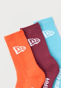 Trzy pary prążkowanych skarpet w kolorze pomarańczowym, burgundowym i jasnoniebieskim. Każda z nich ma biały logo na mankiecie z nazwą marki w blokowych literach.