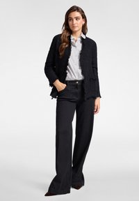 Kobieta o długich brązowych włosach, ubrana w czarną teksturowaną kurtkę, pasiastą koszulę i szerokie czarne spodnie, stoi z jedną ręką w kieszeni.