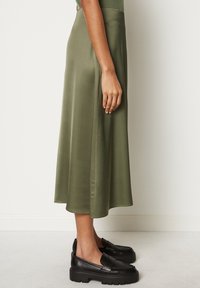 Falda midi de satén verde oliva con una textura suave y un brillo sutil, combinada con unos mocasines negros de suela gruesa y punta redonda con textura.