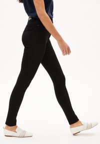 Jeans skinny negros, de cintura alta, con una textura suave. Llevados con zapatos blancos que presentan un patrón tejido y una lengüeta en el talón trasero.