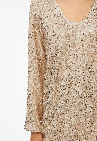 Beżowa sukienka pokryta cekinami z dekoltem w serek i długimi rękawami, z fakturowaną powierzchnią wykonaną z małych, okrągłych cekinów.