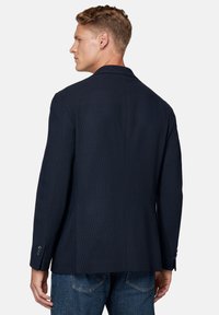 Granatowy blazer z teksturowanej tkaniny, z pojedynczym rozporkiem z tyłu i zapinanymi mankietami, zestawiony z niebieskimi dżinsami, przedstawiony z boku.