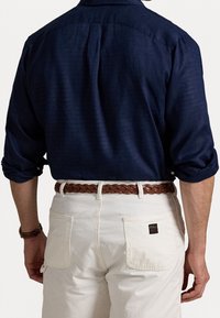 Donkerblauw overhemd met knopen en opgerolde mouwen, gecombineerd met witte broek en een bruine gevlochten riem. Voorzien van een label op de achterzak.