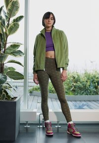 Zelená zipová bunda, fialový crop top, olivové legíny s opakovaným vzorem loga a burgundské tenisky s růžovými akcenty. Vnitřní prostředí s rostlinami.