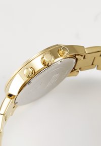 Montre en métal doré avec trois couronnes, boîtier rond lisse et bracelet à maillons. L'arrière présente une surface argentée avec des détails gravés.