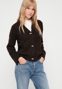 Cardigã castanho de malha com decote em V, fecho de botões e dois bolsos frontais. Usado sobre uma blusa branca e combinado com jeans azul claro.