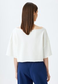Hvid, cropped top med brede ærmer og rå kant. Glat tekstur, åben rygdesign, båret med mørkeblå bukser.